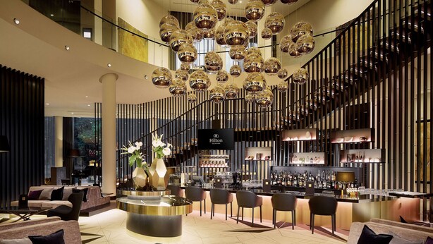 A modern hotel lobby with a bar area, seating arrangements, and a staircase. - Hilton Tallinn Park (Tallinn)