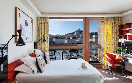 A hotel room with a bed, a desk, a chair, a window with a city view, and a plant.