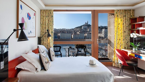 A hotel room with a bed, a desk, a chair, a window with a city view, and a plant.
