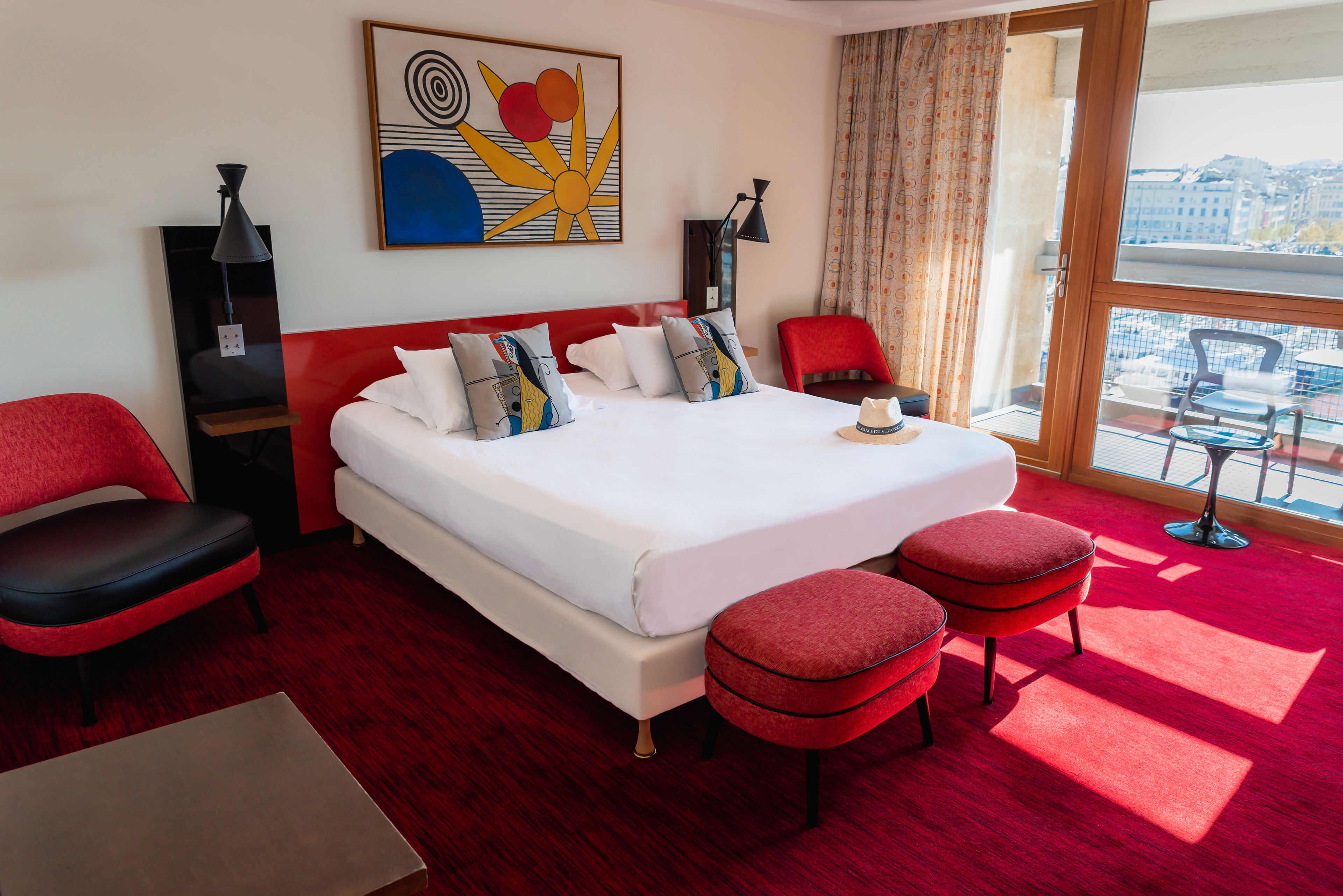 Ein Hotelzimmer mit einem großen Bett, roten Sitzmöbeln, einem kleinen Tisch und einem großen Fenster mit Blick auf die Stadt.