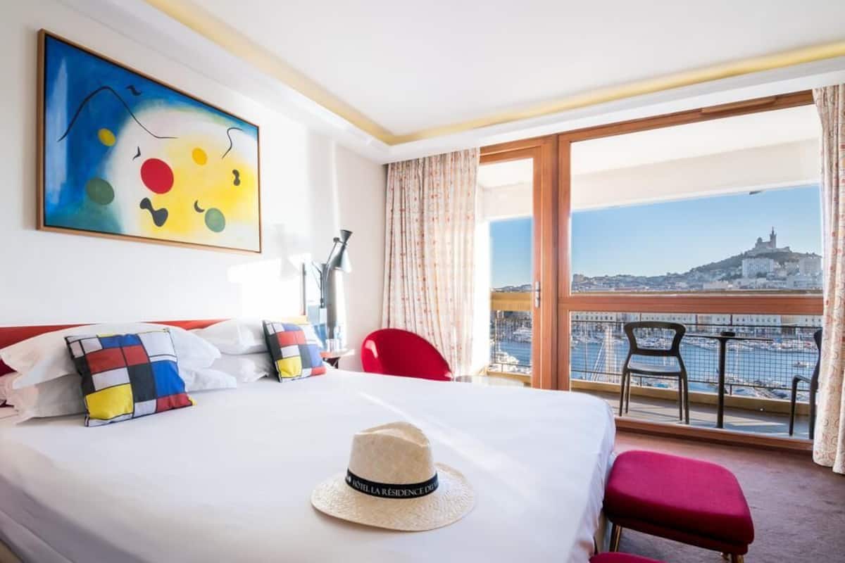 A hotel room with a bed, a hat, a painting, a view of a cityscape, and a balcony with chairs.