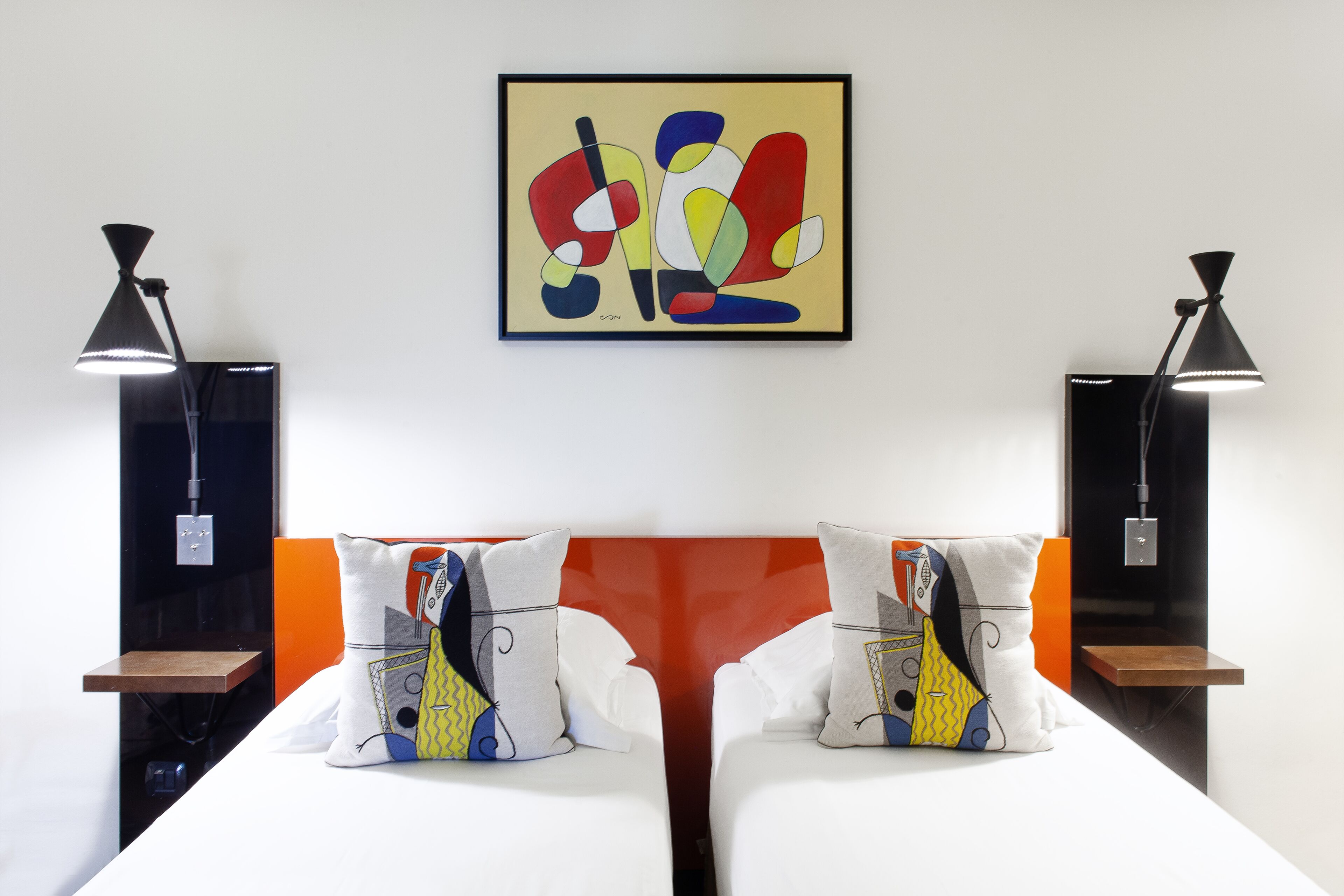 Habitación de hotel con dos camas, cada una con un almohadón de diseño geométrico. Hay dos mesitas de noche, cada una con una lámpara. Un cuadro abstracto y colorido cuelga de la pared sobre las camas.