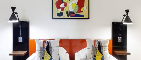 Habitación de hotel con dos camas, cada una con un almohadón de diseño geométrico. Hay dos mesitas de noche, cada una con una lámpara. Un cuadro abstracto y colorido cuelga de la pared sobre las camas.