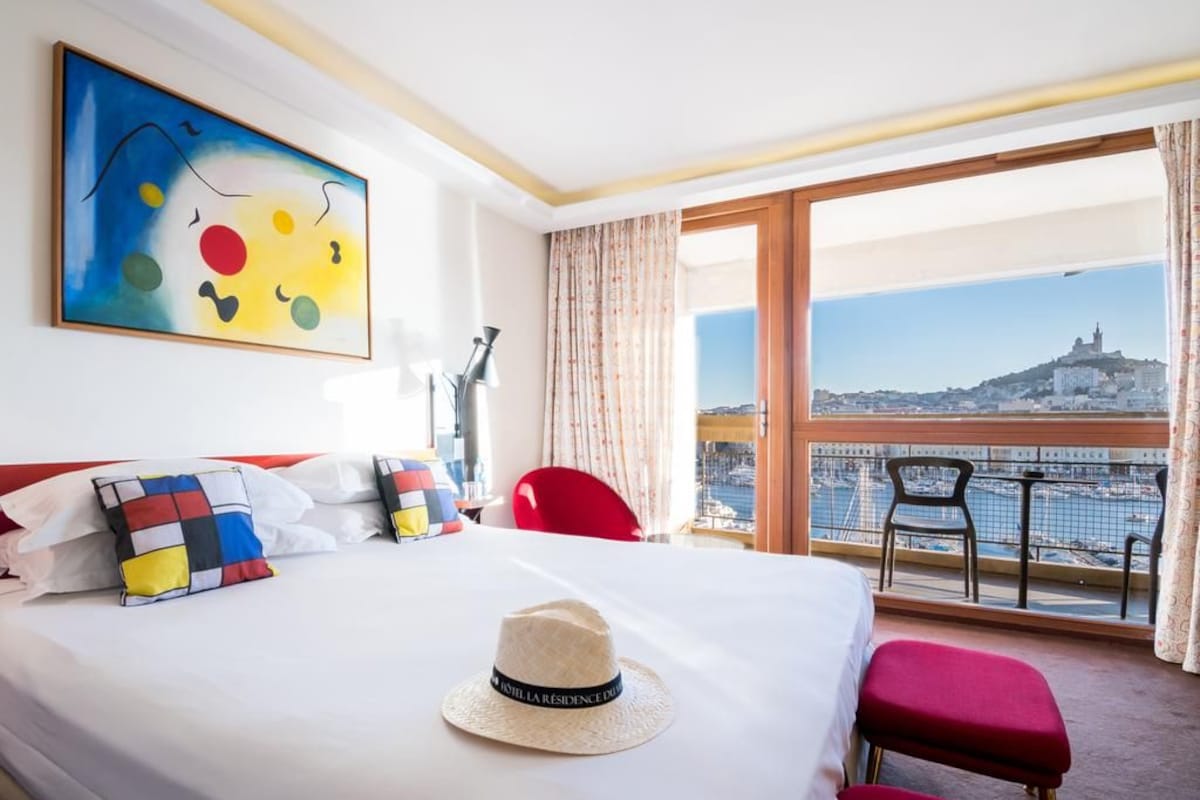 A hotel room with a bed, a hat, a painting, a view of a cityscape, and a balcony with chairs.