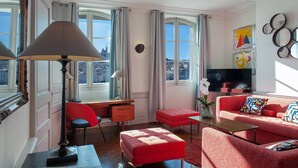 A modern living room with a red sofa, a desk, and a lamp. There are large windows with curtains, a view of the city, and a wall with decorative mirrors.