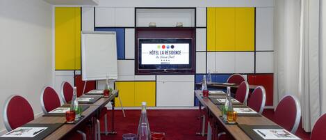 A conference room with a large table, red chairs, a whiteboard, and a TV screen displaying a hotel sign.