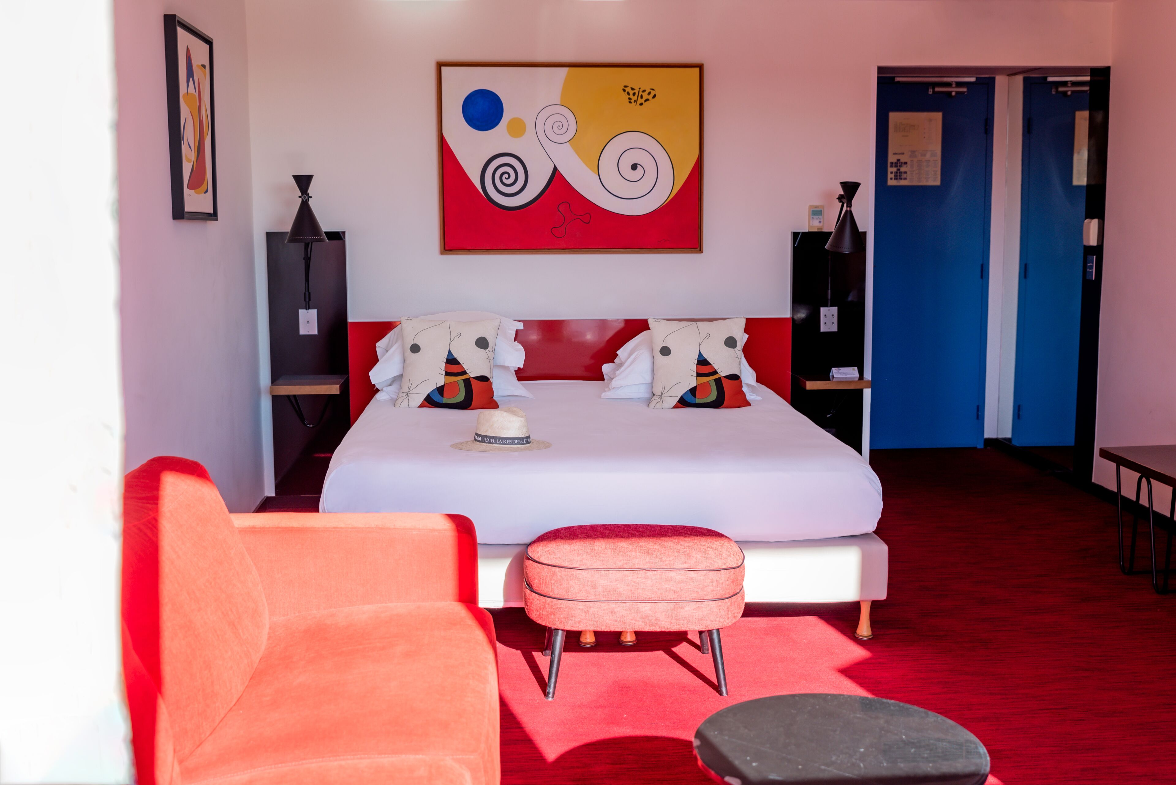 Una habitación de hotel moderna con una cama grande, ropa de cama roja y blanca, una alfombra roja, una silla roja, un pequeño reposapiés y un cuadro en la pared.