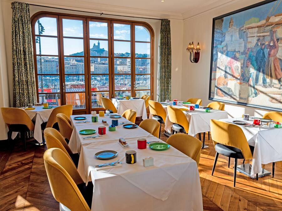 A dining area with tables set for a meal, large windows offering a city view, and a painting on the wall.