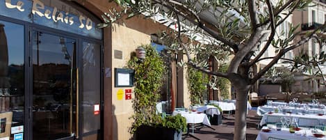 Ein Restaurant mit Außenbereich, Tischen zum Essen, ein großer Baum im Vordergrund und ein Gebäude mit der Aufschrift 'Le Relais 50'.