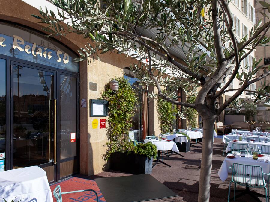 An outdoor restaurant with tables set for dining, a large tree in the foreground, and a building with a sign reading "Le Relais 50".
