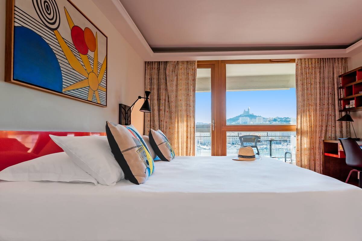 A hotel room with a large bed, a view of a cityscape, and a painting on the wall.