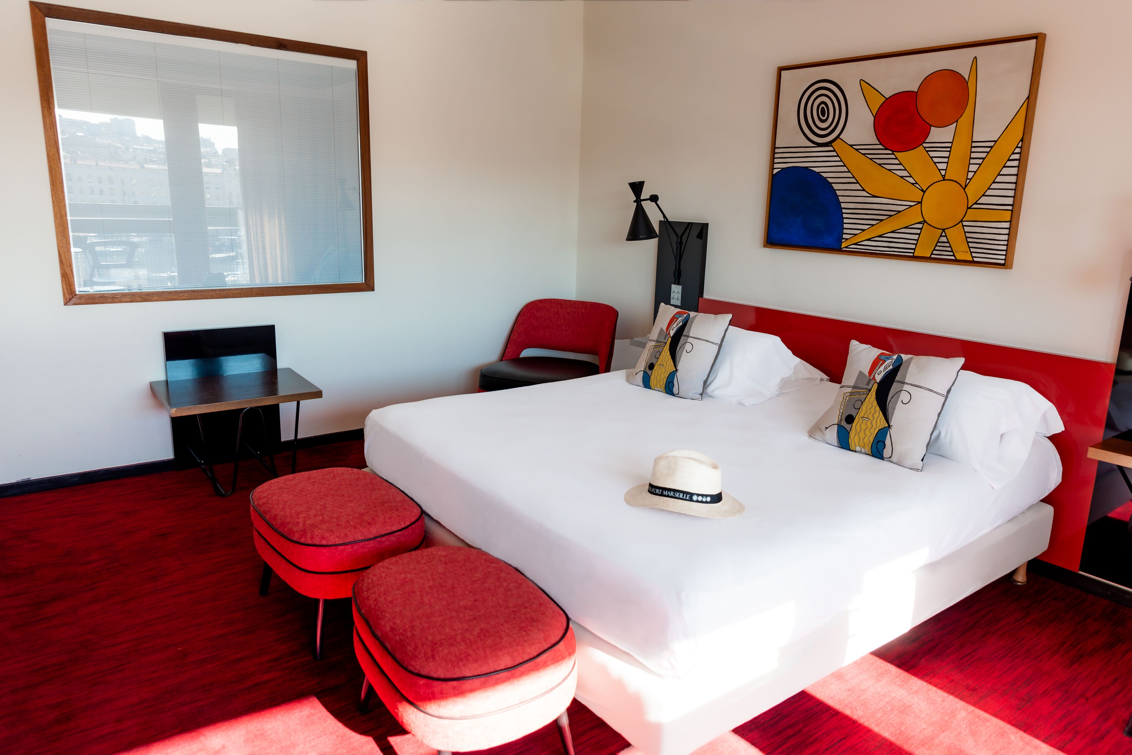 A hotel room with a large bed, a desk, a chair, and a red ottoman.