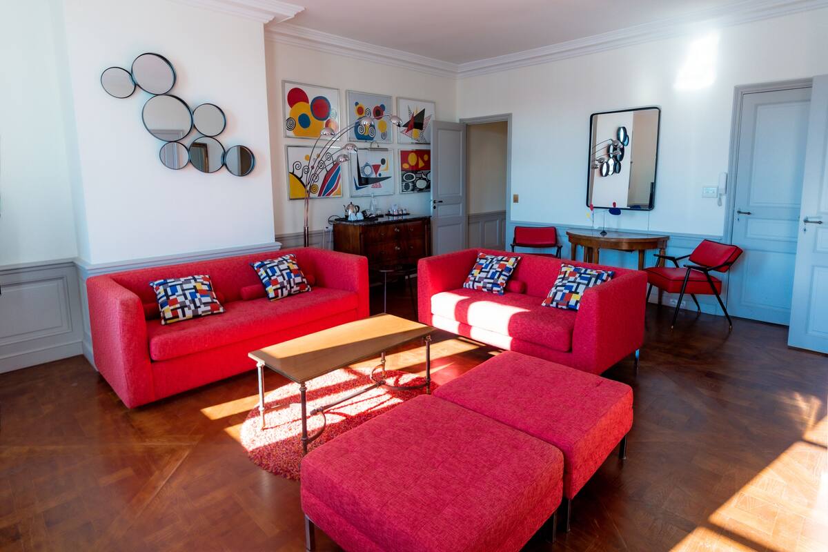 A living room with red sofas and a coffee table, a dining area with a table and chairs, and a door leading to another room.
