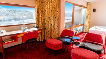 A hotel room with a desk, chairs, a small table, a sofa, and a view of a cityscape through large windows.