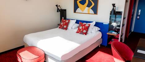 A modern hotel room with a large bed, red and white pillows, a red ottoman, a red chair, a painting on the wall, and a water dispenser.