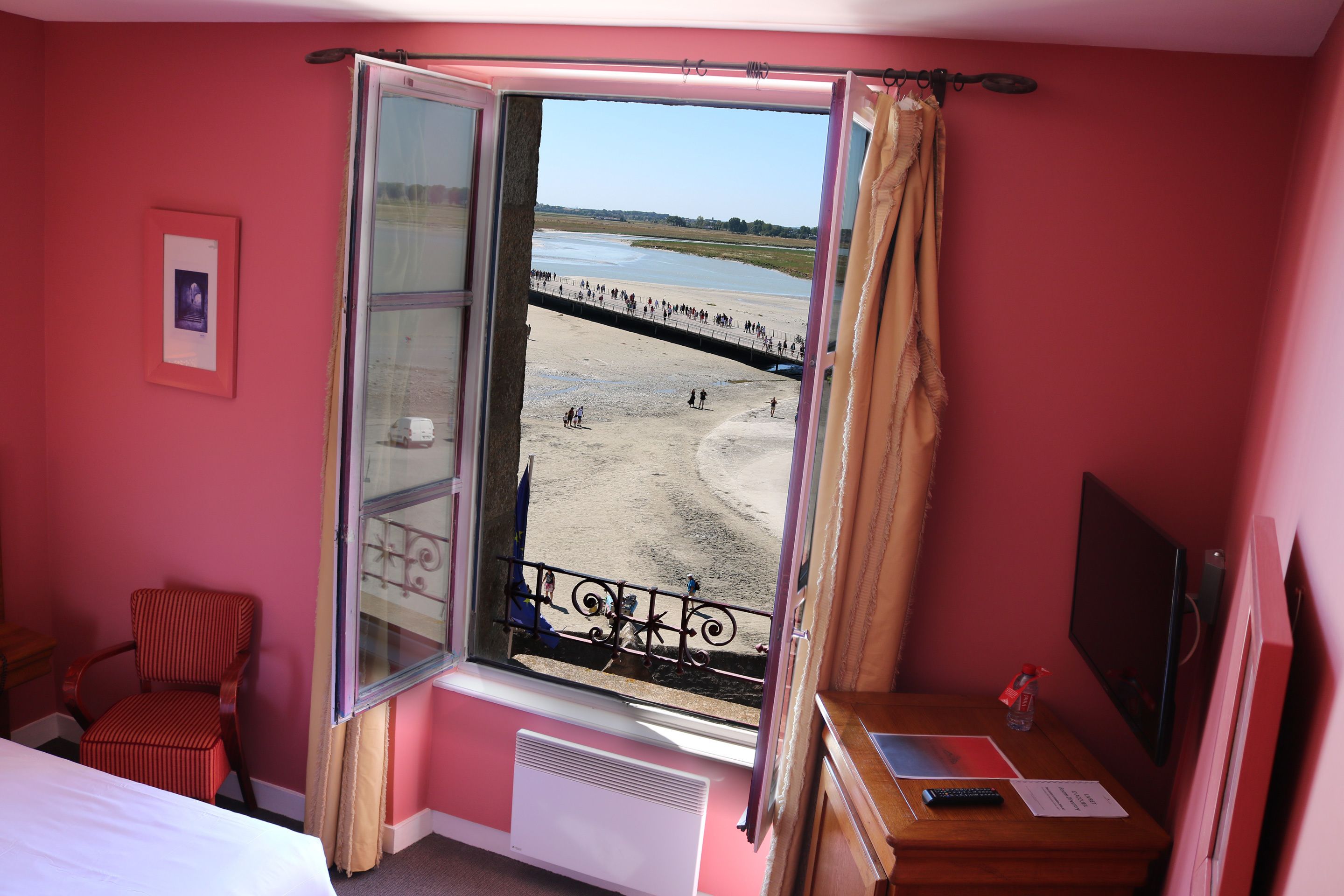 Een kamer met een groot raam met uitzicht op een strand, een bed, een stoel en een bureau met een computer en papieren.