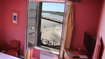 Een kamer met een groot raam met uitzicht op een strand, een bed, een stoel en een bureau met een computer en papieren.