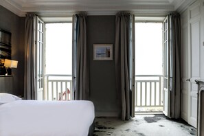 A hotel room with a large bed, a window with curtains, a framed picture on the wall, and a balcony with a railing.