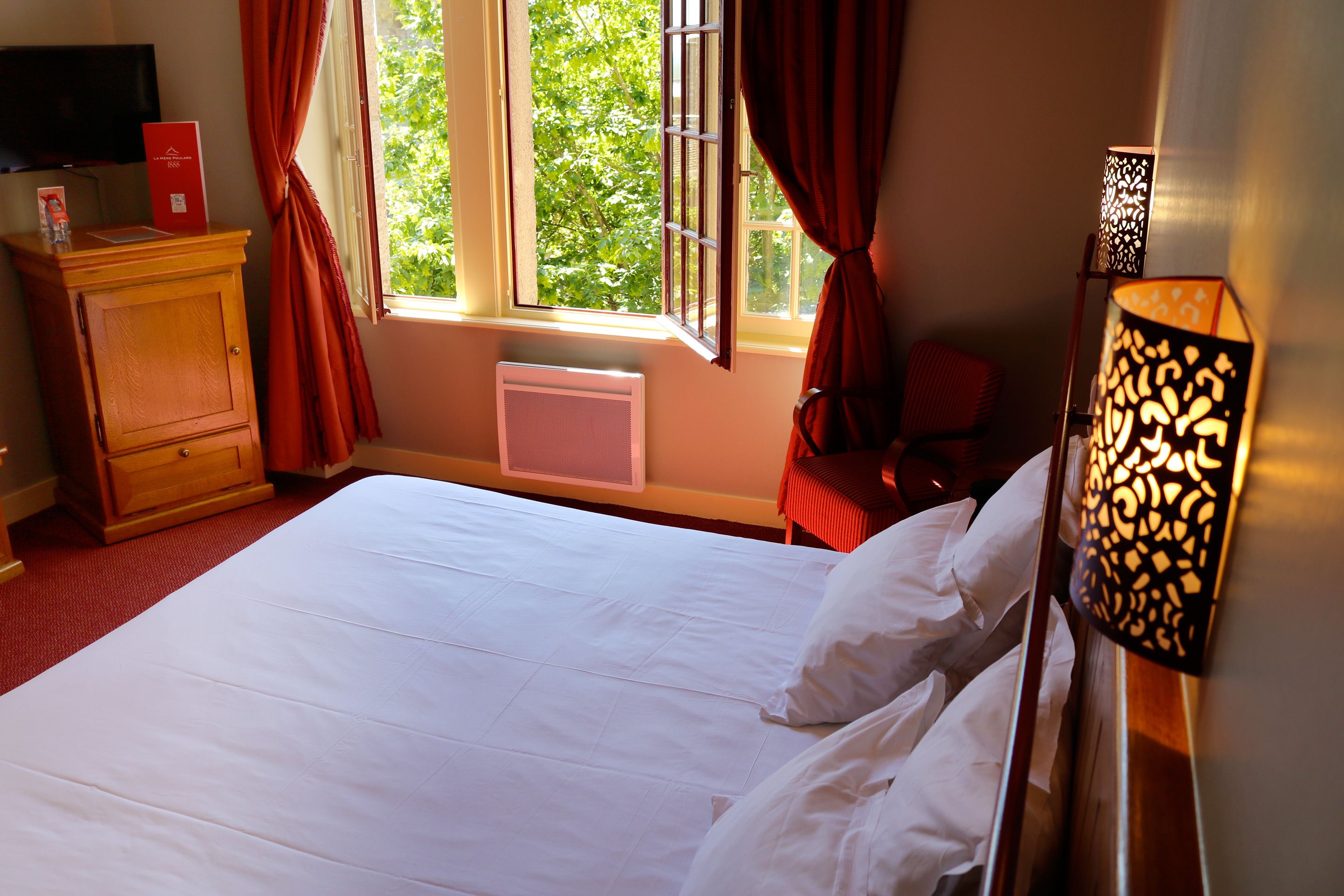 Une chambre d’hôtel avec un grand lit, une table de chevet en bois, une chaise rouge, un téléviseur et une fenêtre avec des rideaux.