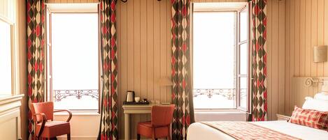 A hotel room with a bed, a desk with a chair, and a window with curtains.