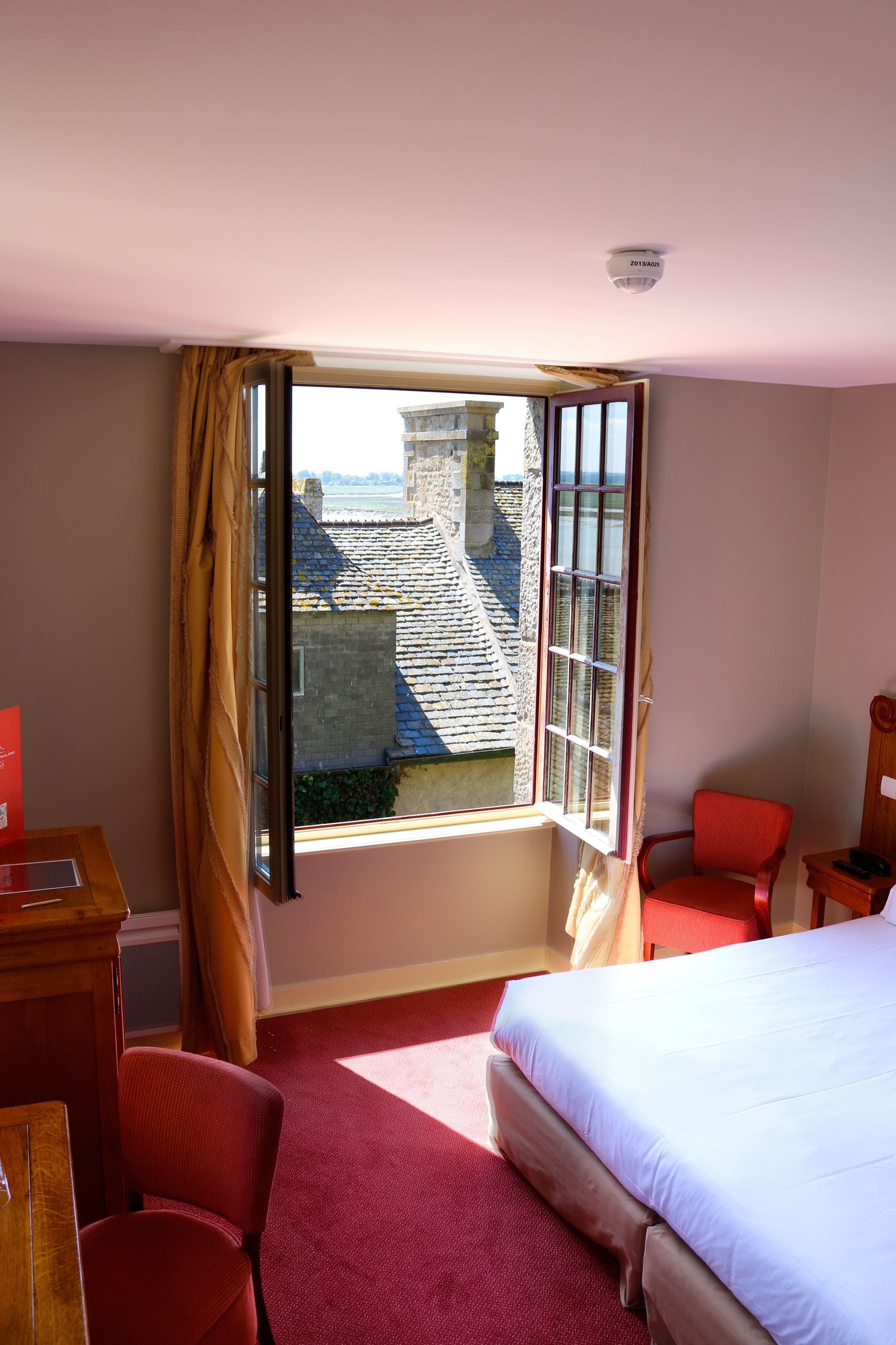A hotel room with a large bed, a desk with a chair, a window with curtains, and a view of buildings outside.