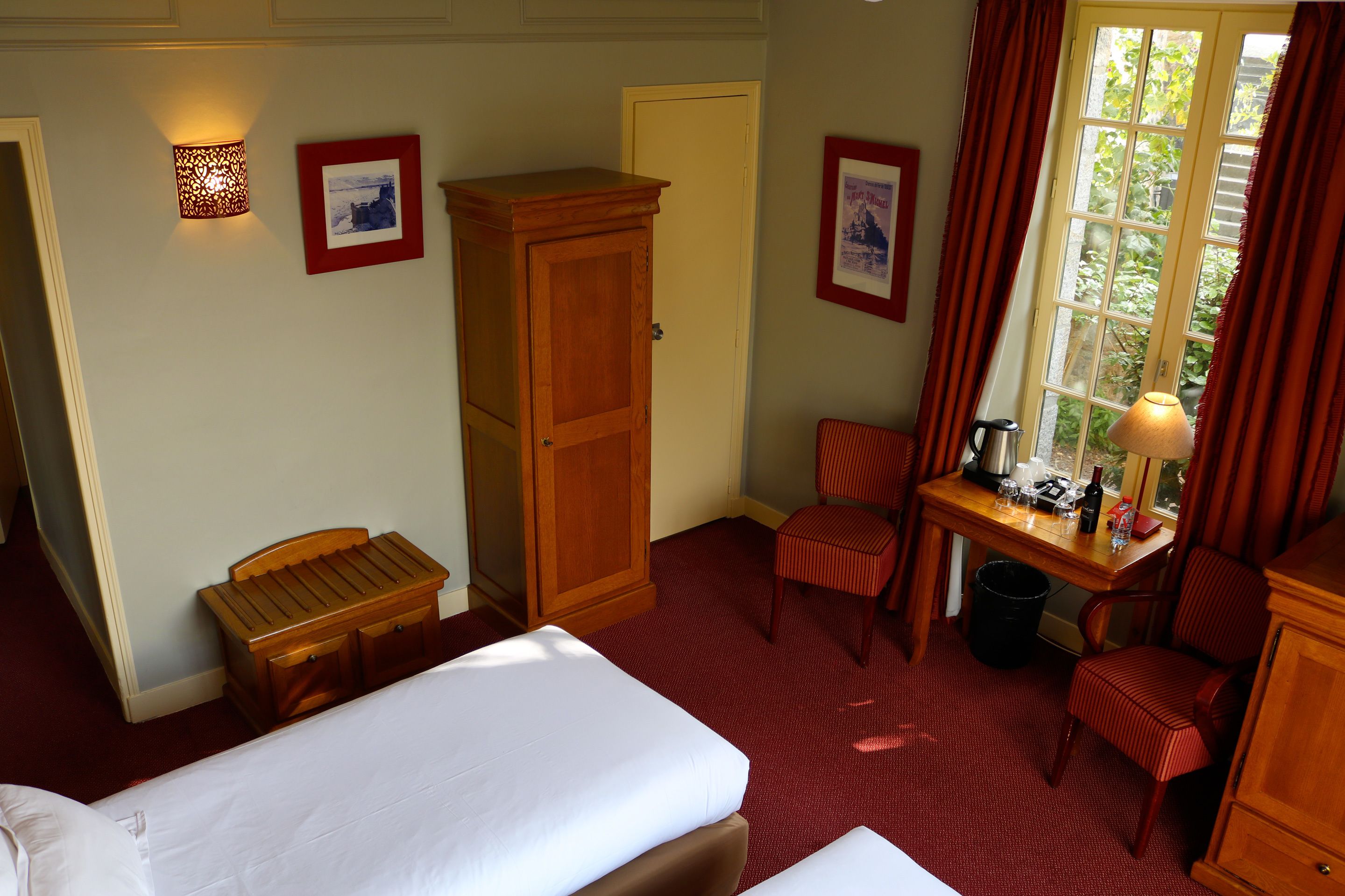 A hotel room with a bed, a desk with a lamp, a chair, a wardrobe, and a window with curtains.