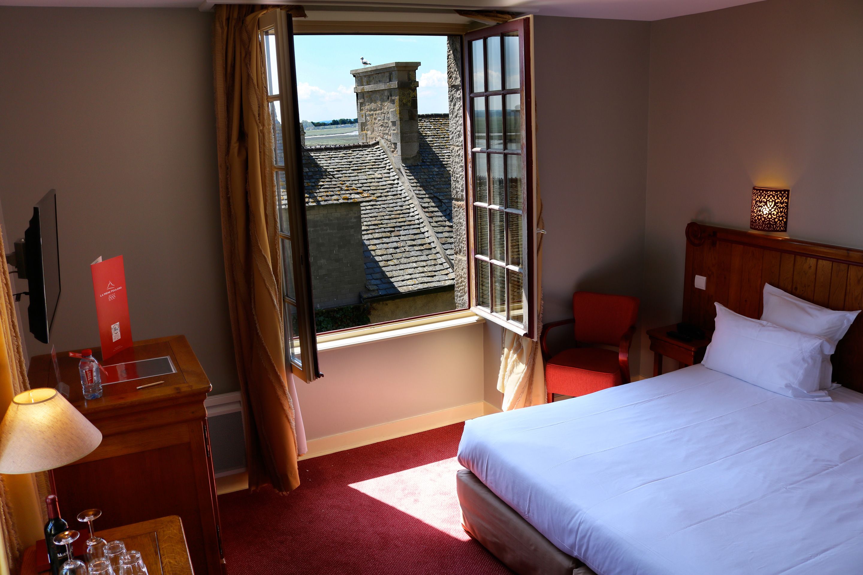 A hotel room with a bed, a desk with a lamp, a chair, a window with curtains, and a view of a stone building outside.