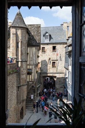 A narrow medieval street with stone buildings, a tower, and a crowd of people.