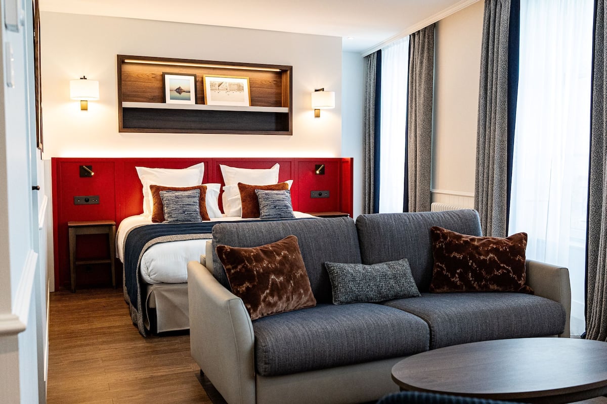Una habitación de hotel moderna con una cabecera roja, un sofá gris y una mesita de noche de madera.