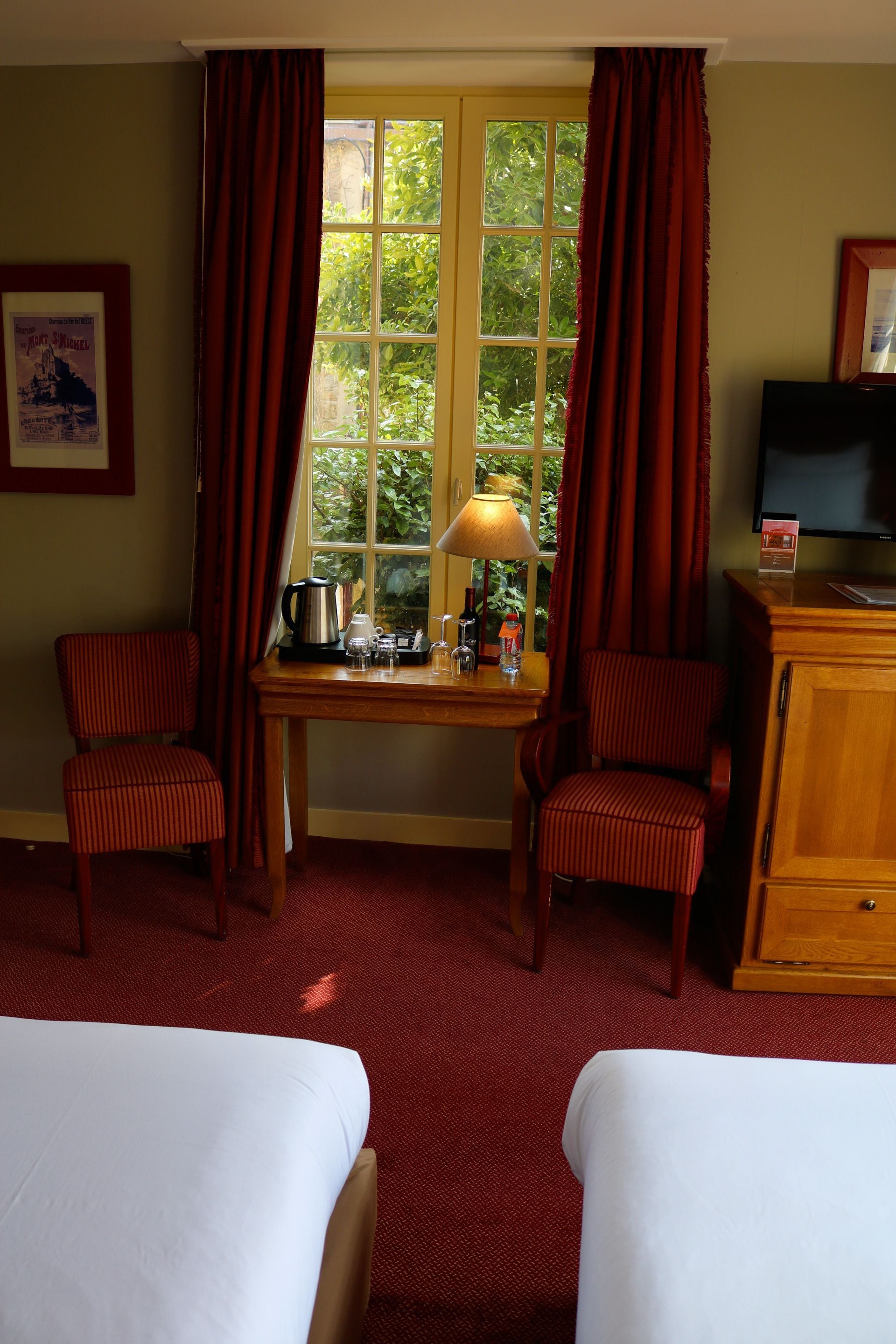 A hotel room with two single beds, a wooden desk with a lamp, a television, and a window with curtains.