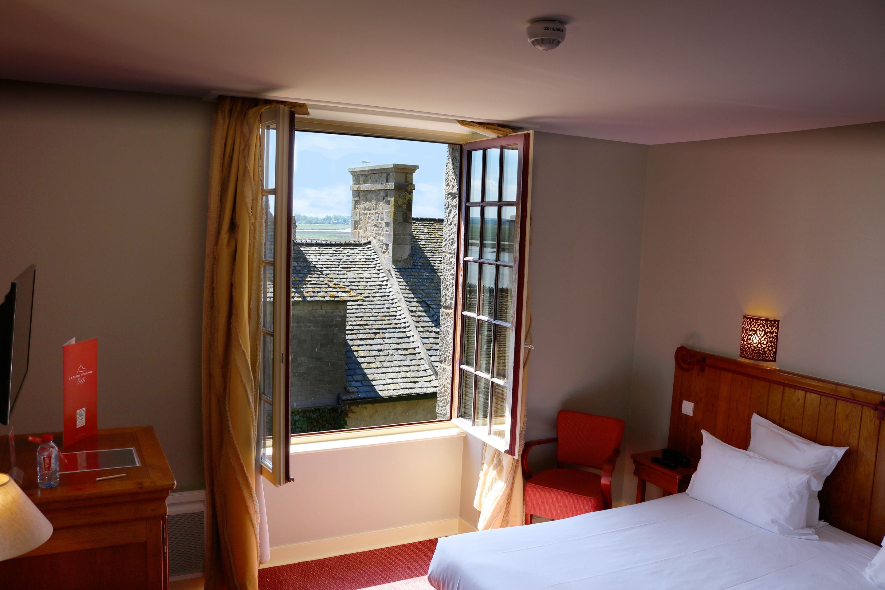 A hotel room with a bed, a desk, a chair, a nightstand, a window with curtains, and a view of the outside.