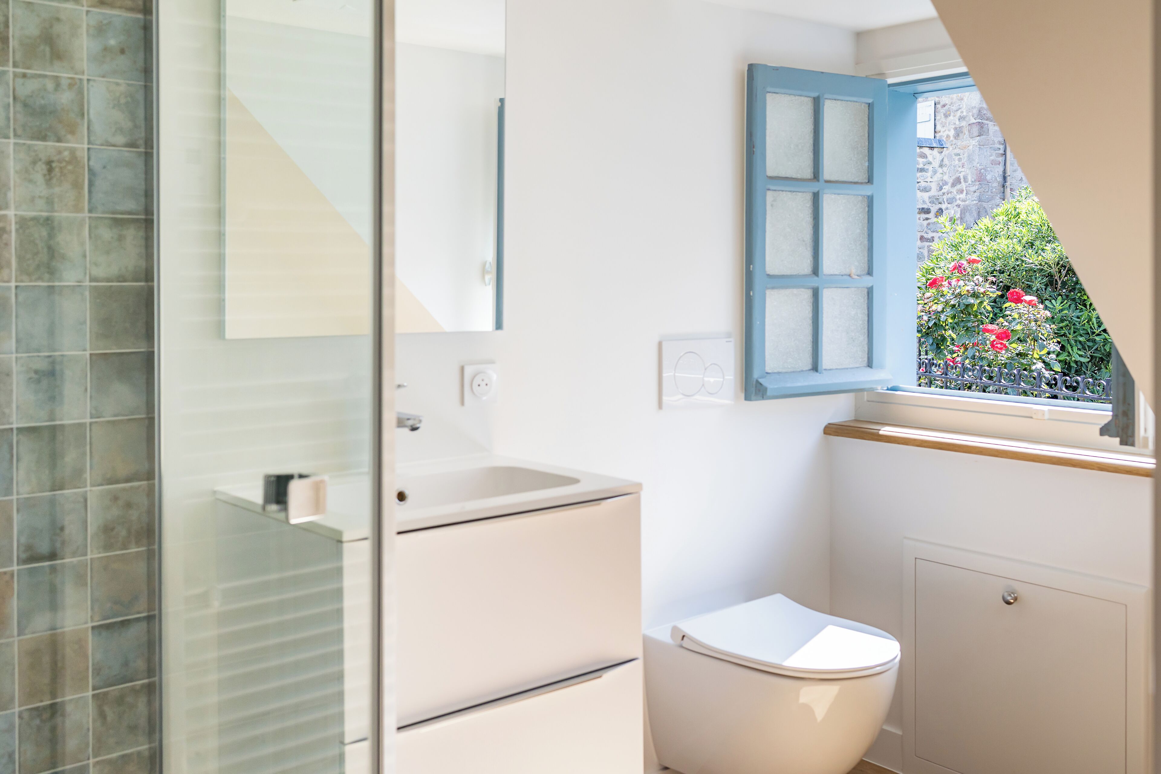Une salle de bain avec une douche à parois de verre, un lavabo blanc, des toilettes et une fenêtre avec des volets bleus.