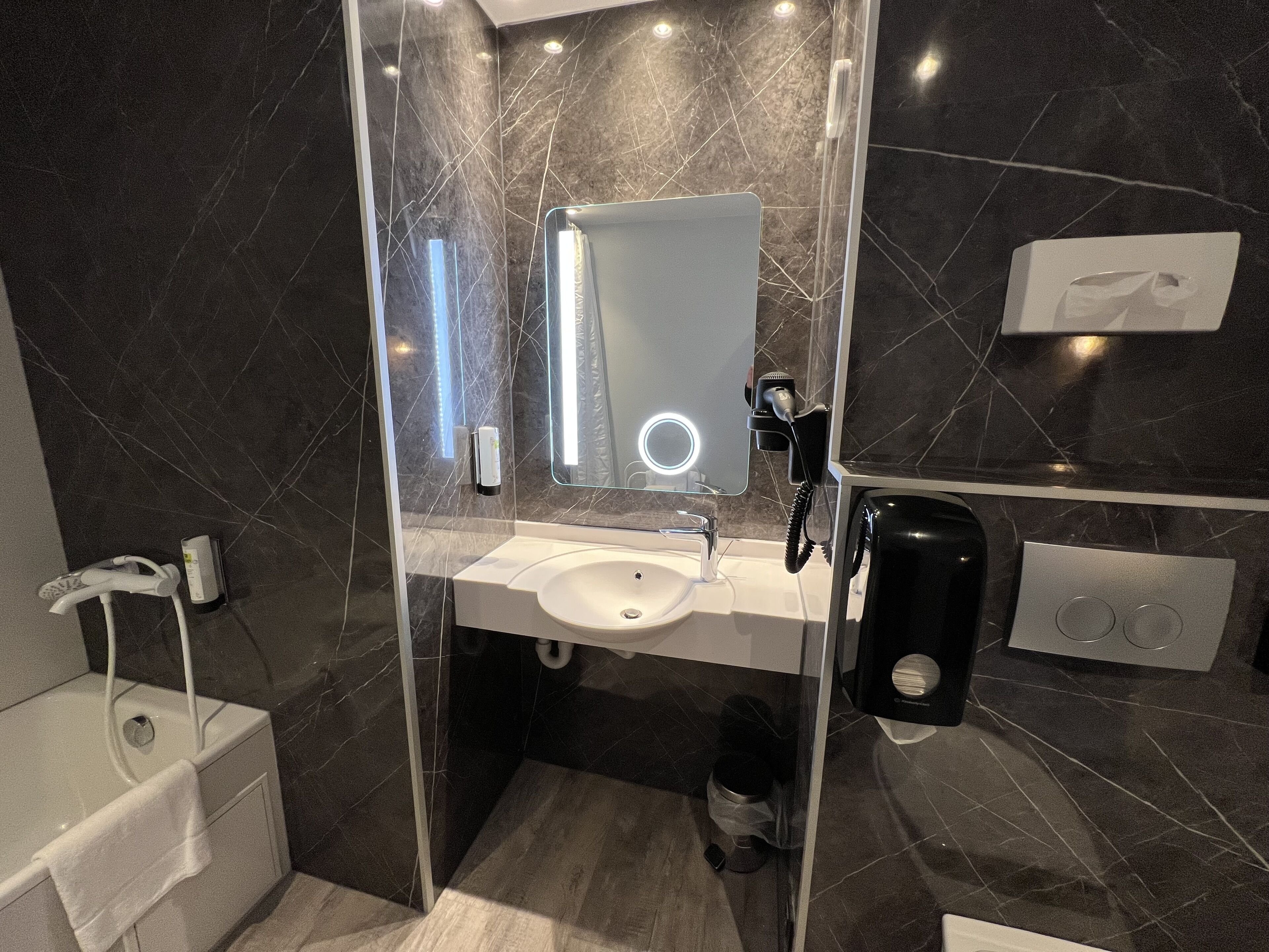 A modern bathroom with a glass-enclosed shower, a white sink, a mirror with built-in lighting, and a handheld hair dryer.