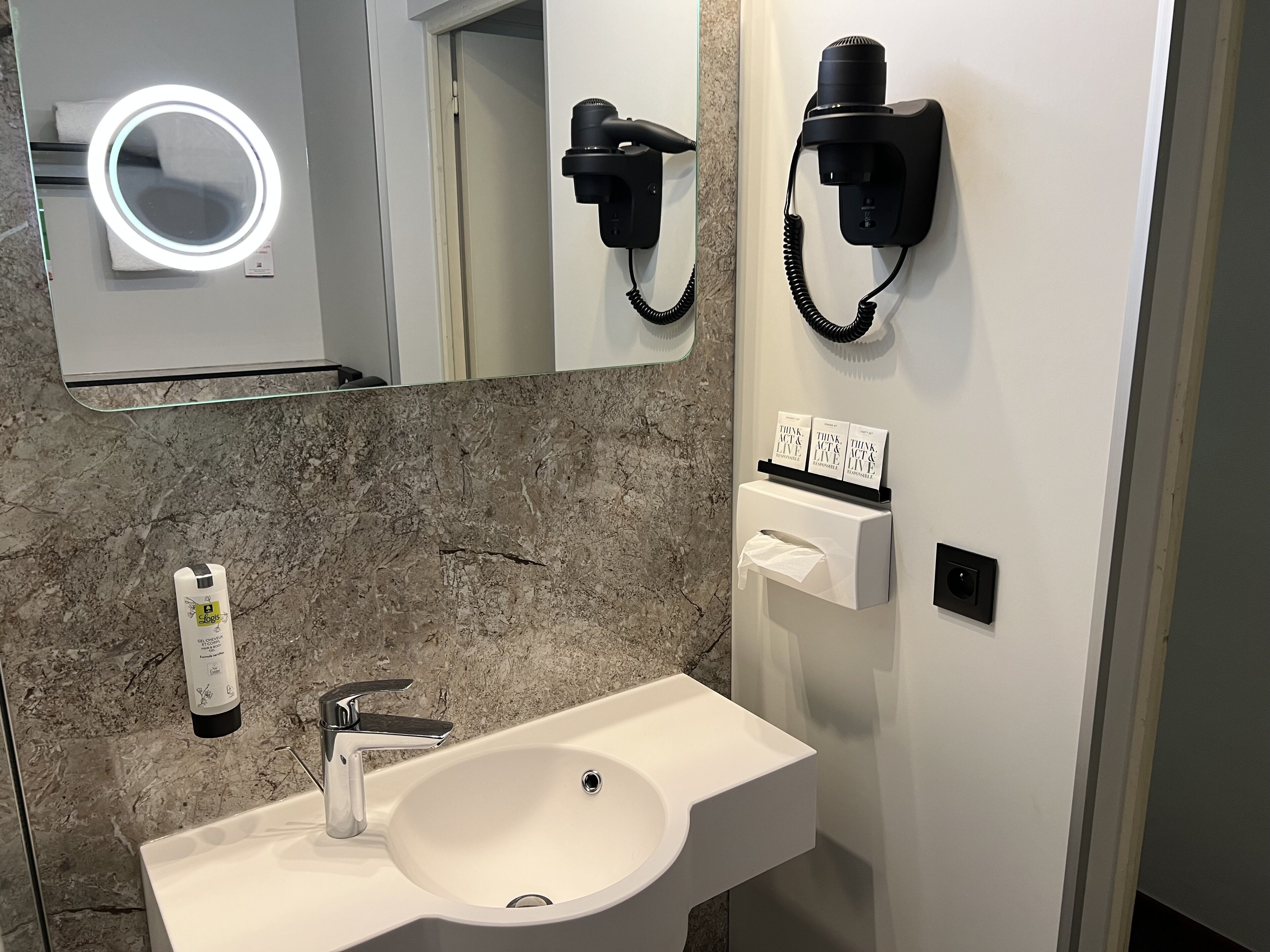 A bathroom with a white sink, a mirror with a ring light, a hairdryer, and a tissue dispenser.