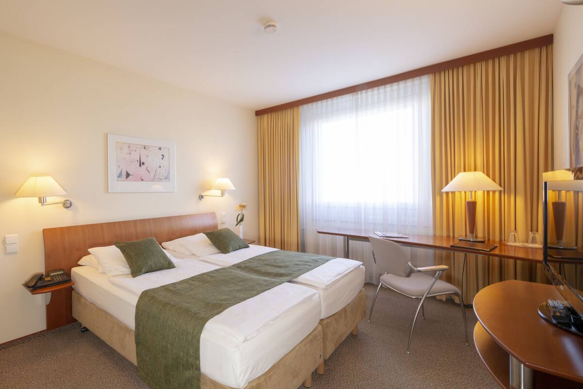 Habitación de hotel con cama doble, escritorio, silla y ventana grande con cortinas.