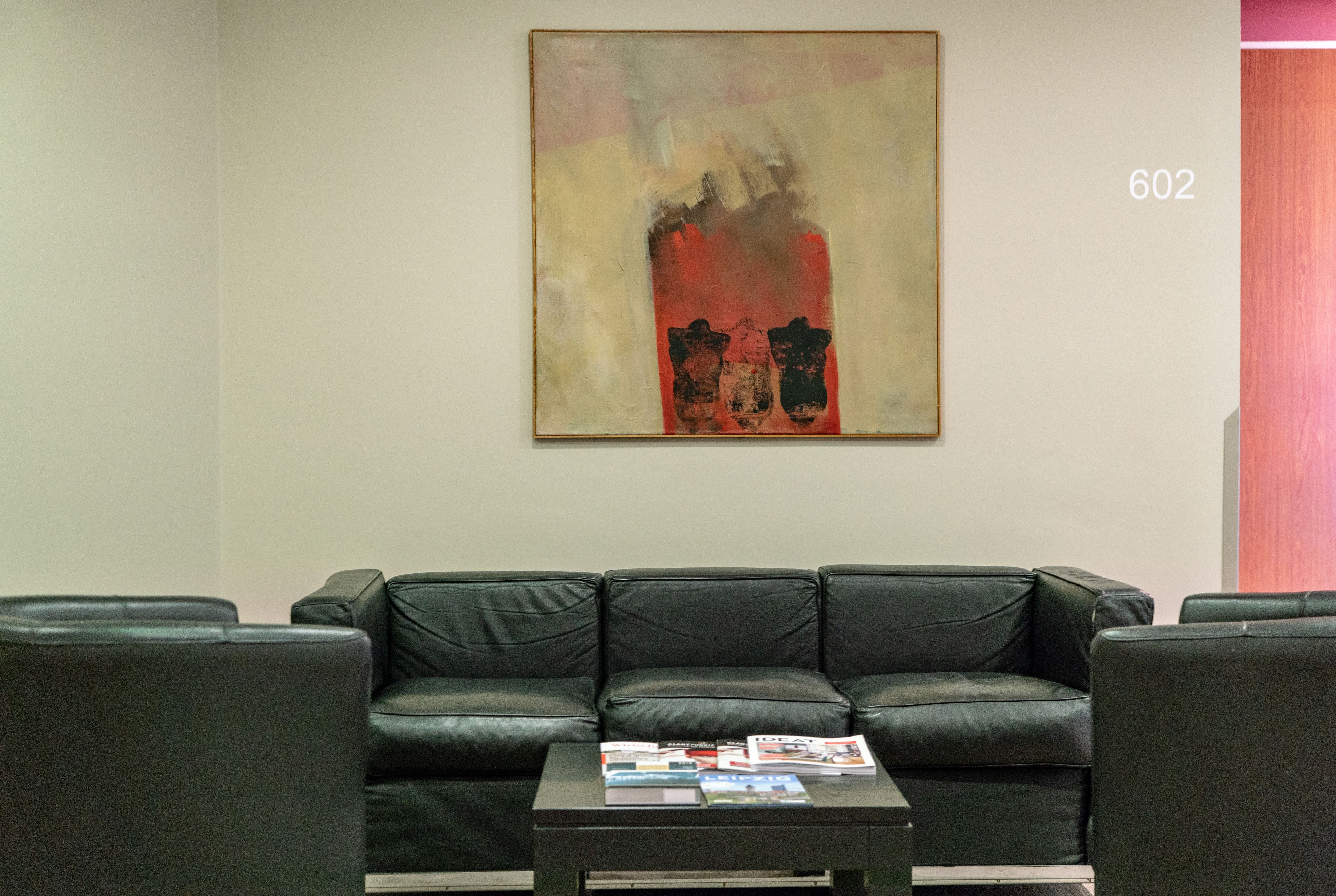 A modern living room with a black leather sofa, a coffee table with magazines, and a large abstract painting on the wall.