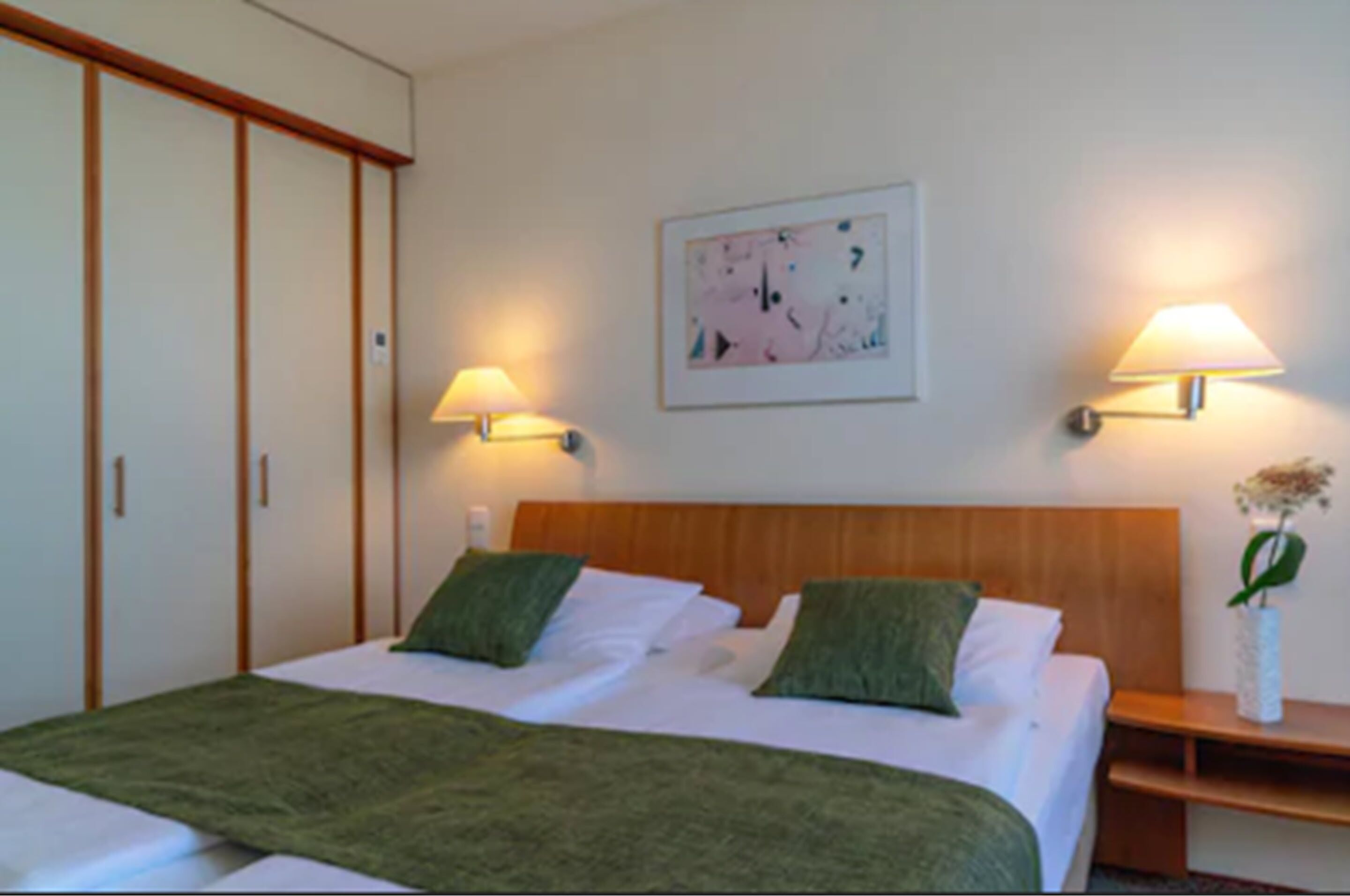 Ein Hotelzimmer mit einem Bett, zwei Nachttischlampen, einem Nachttisch, einer Vase mit Blumen und einem Bild an der Wand.