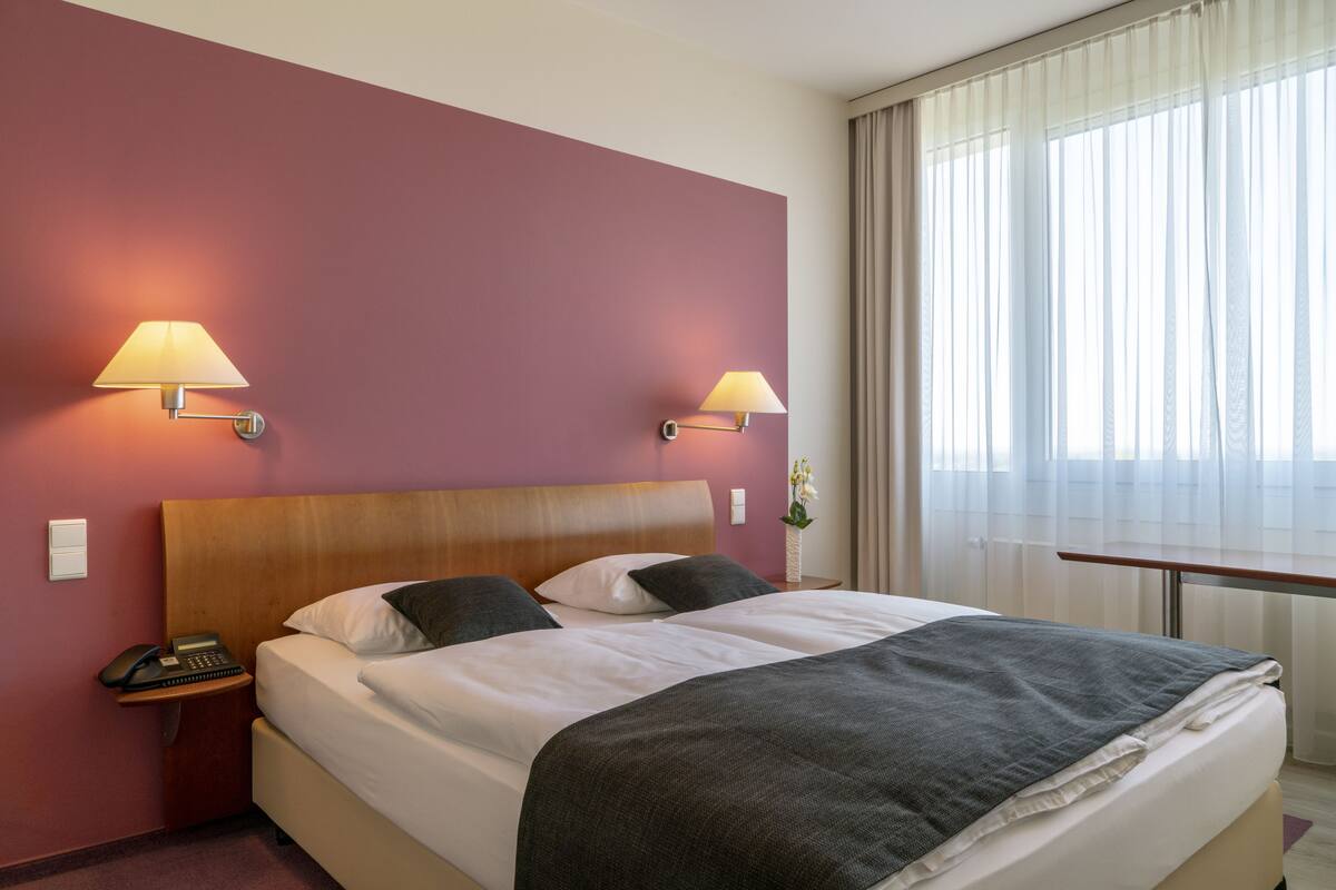 Una habitación de hotel con una cama grande, dos lámparas de noche, una mesita de noche con teléfono y una ventana con cortinas translúcidas.