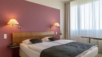 Ein Hotelzimmer mit einem großen Bett, zwei Nachttischlampen, einem Nachttisch mit Telefon und einem Fenster mit durchsichtigen Vorhängen.