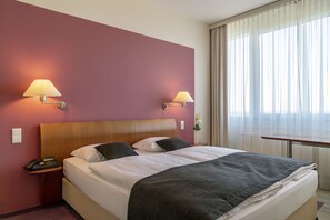 Quarto de hotel com cama grande, duas luminárias de cabeceira, mesa de apoio com telefone e janela com cortinas leves.