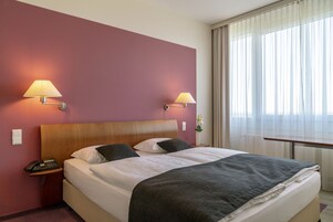 Una habitación de hotel con una cama grande, dos lámparas de noche, una mesita de noche con teléfono y una ventana con cortinas translúcidas.