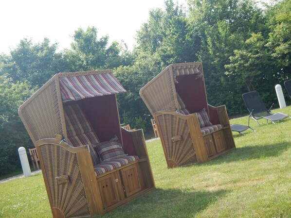 Two wicker daybeds with striped cushions and a red canopy, placed on a grassy lawn with trees in the background.