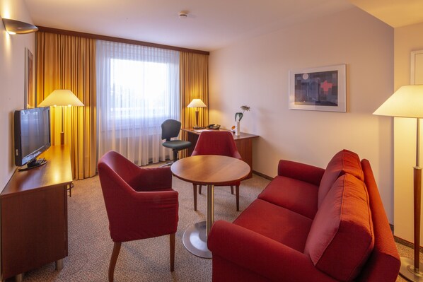 A hotel room with a red sofa, a small round table, a TV, and a desk with a lamp.
