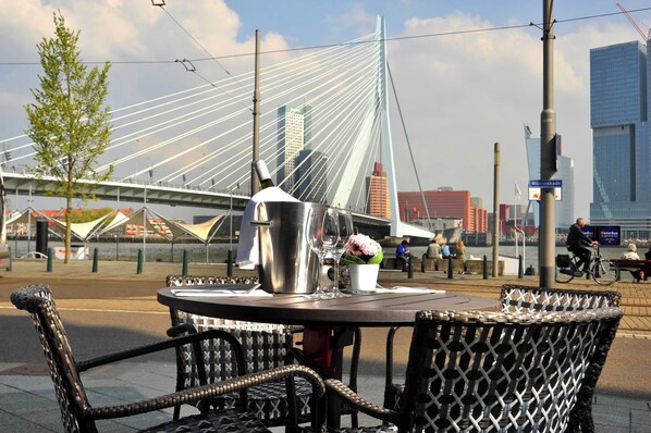 An outdoor seating area with a table and chairs, a metal coffee pot, a glass, and a flower arrangement. In the background, there is a cable-stayed bridge and modern buildings. - Thon Hotel Rotterdam (Rotterdam)