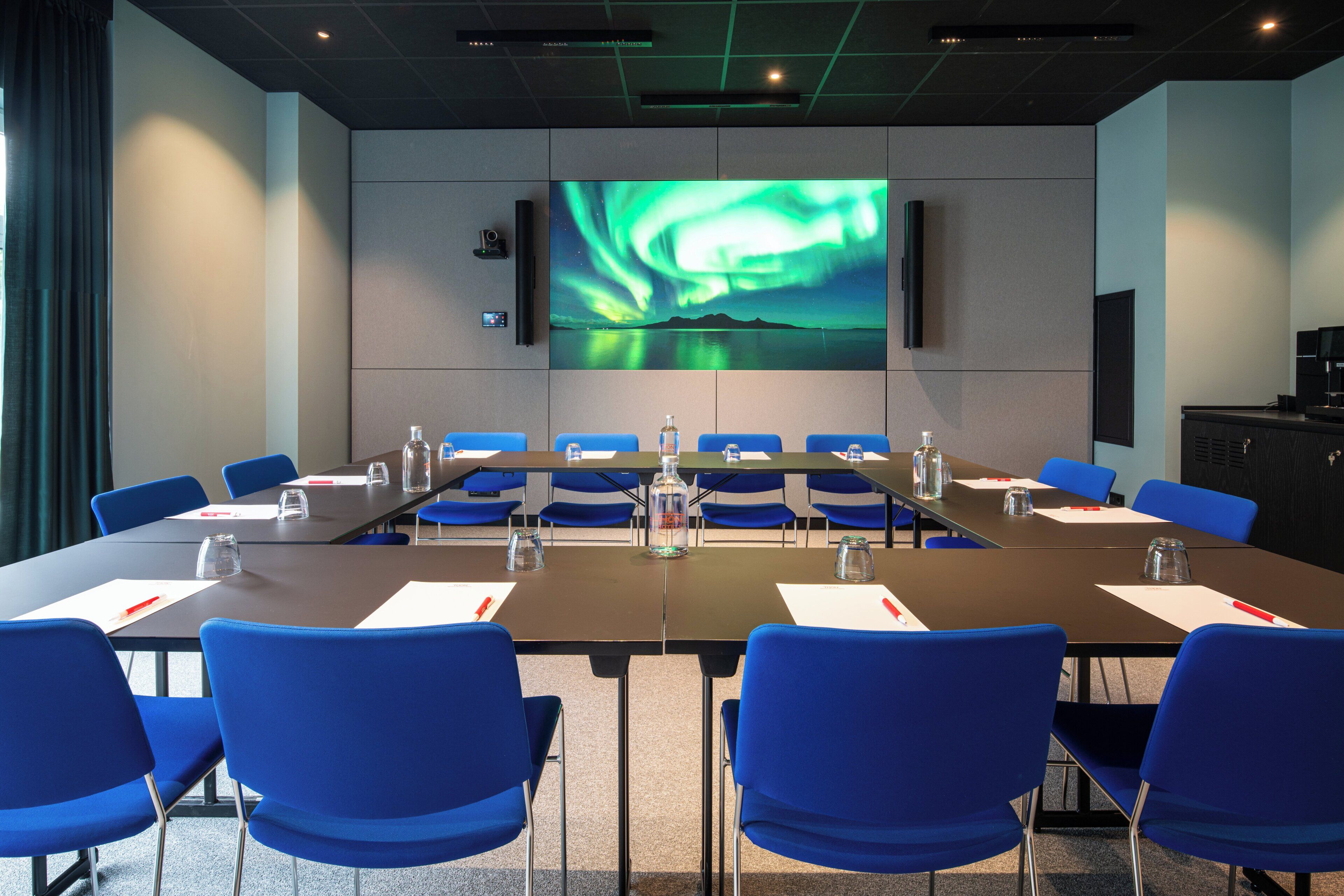 A conference room with a large screen displaying a nature scene, blue chairs, and a long table with water bottles and nameplates.