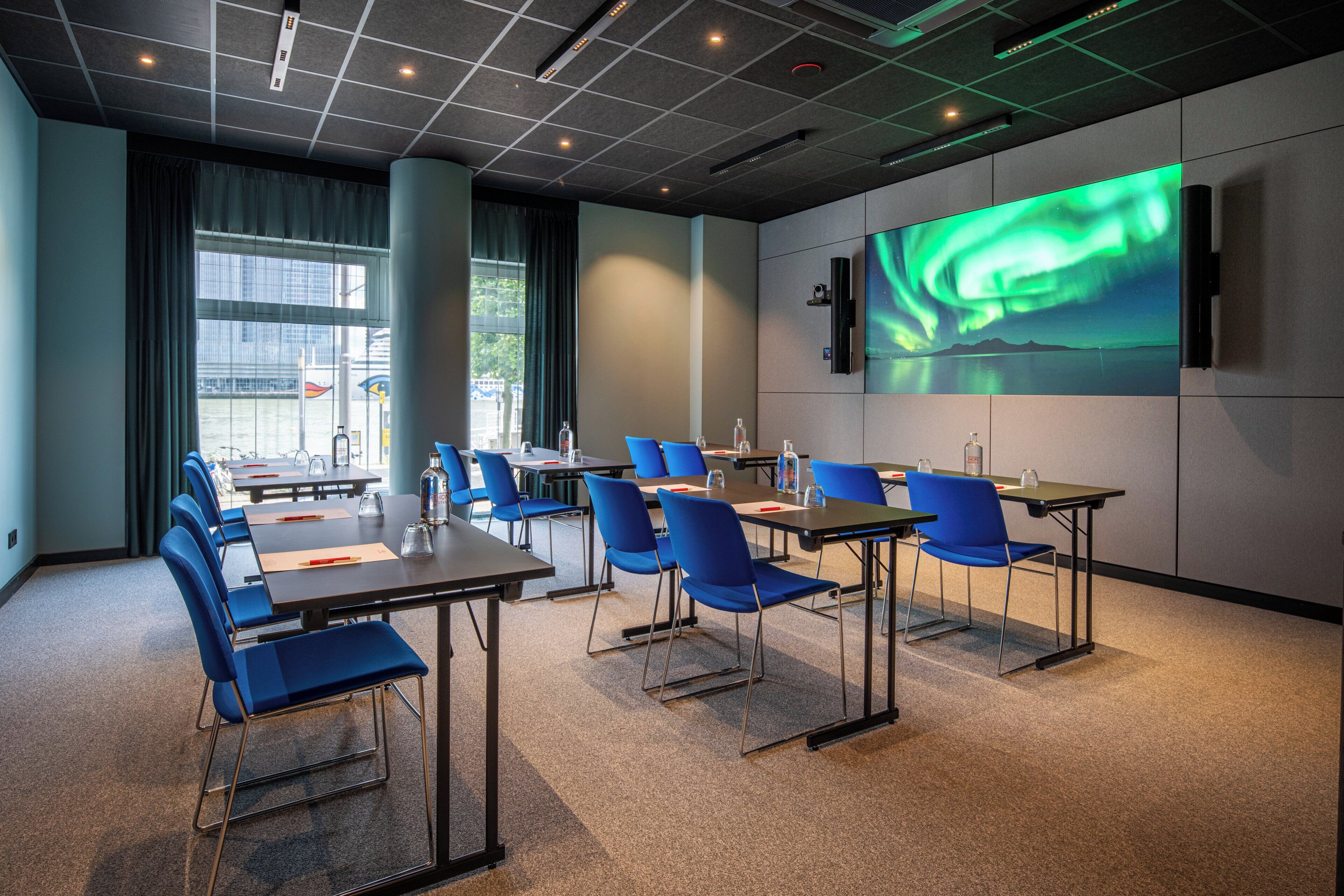 Un salón de conferencias con mesas, sillas y una pantalla grande que muestra una imagen abstracta verde.