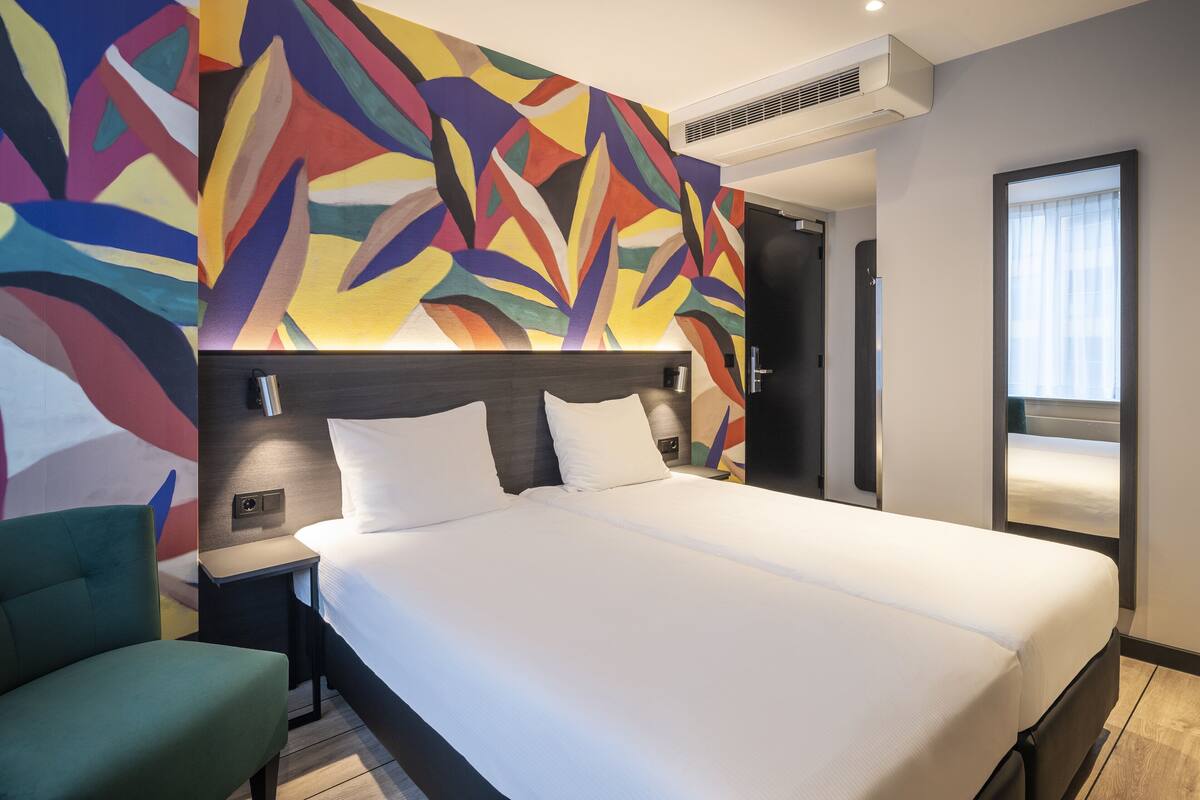 Een moderne hotelkamer met een groot bed, een groene fauteuil, een kleurrijk abstract schilderij en een raam met gordijnen.