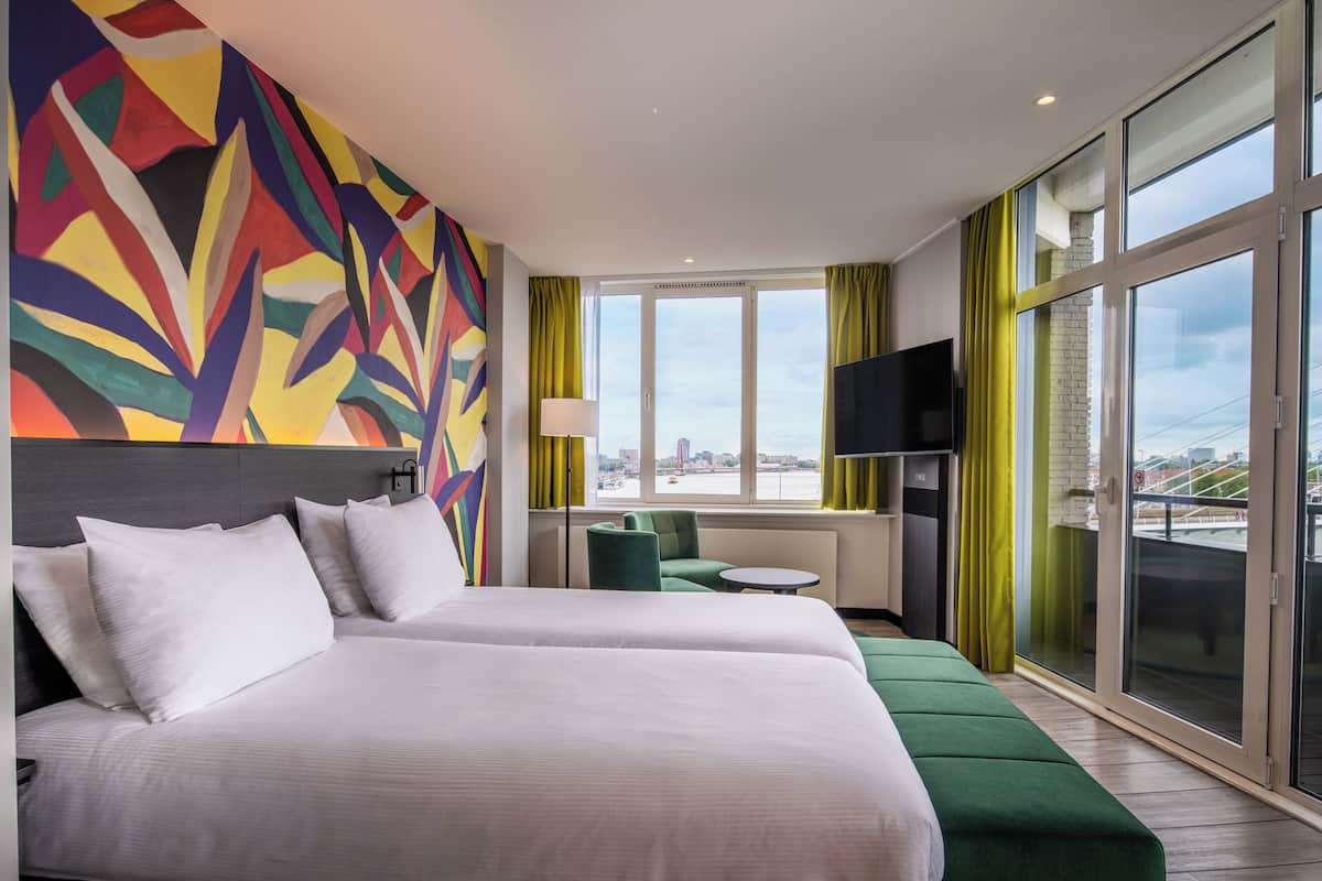 Een hotelkamer met twee bedden, een kleurrijk abstract schilderij, een televisie, een raam met uitzicht op een stadsgezicht en een klein tafeltje.