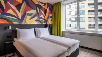 A hotel room with a large bed, a nightstand, a colorful abstract wall, a window with curtains, and a view of a building.