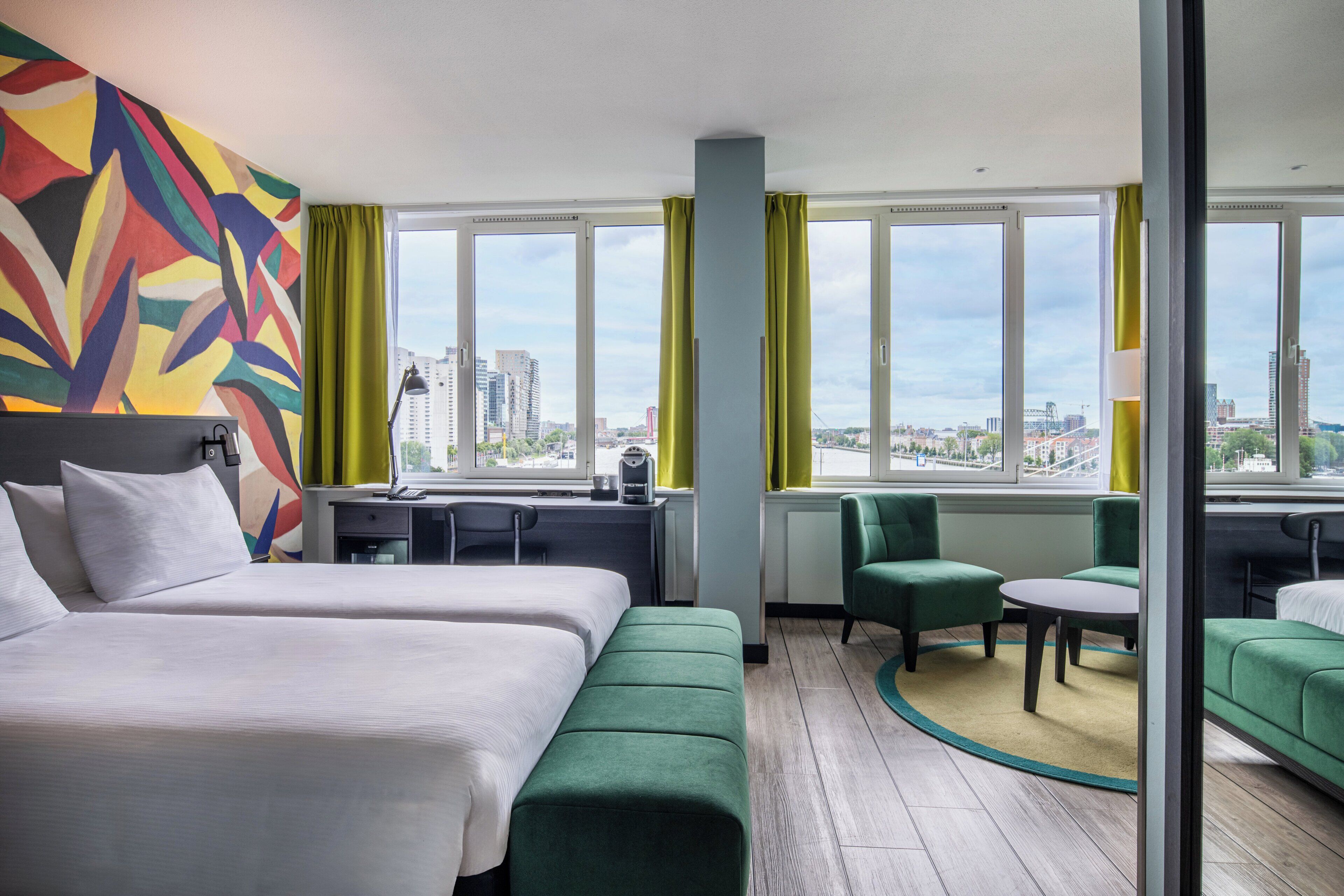 A hotel room with two beds, a desk, a chair, a small table, and a view of the cityscape.
